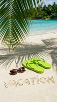 Tropical beach scene with green flip flops, sunglasses, and vacation written in sand under palm leaves with turquoise sea in background.