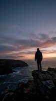 Silhouetted male hiker standing on rocky cliff overlooking ocean at sunset, wearing backpack, dramatic sky with orange and purple clouds, coastal landscape, adventure and exploration theme.