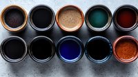Colorful mineral pigments and natural dyes in ceramic bowls arranged in two rows, showing vibrant hues of blue, green, red, and black on gray background.