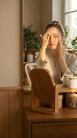 Young woman of East Asian descent applying skincare product to face while sitting at wooden desk with mirror, plants, and cosmetic items in bright natural light from window.