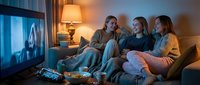Three young adults of mixed ethnicities sitting on a couch watching television together in a cozy living room with warm lighting and snacks, ideal for entertainment and lifestyle content.