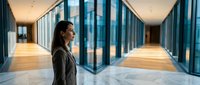 Professional Asian businesswoman in suit standing in modern corporate office hallway with glass walls and marble floors during evening hours.