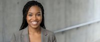 Professional African American businesswoman with braided hair smiling confidently in modern office environment wearing gray blazer for corporate presentations.