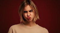 Young woman with blonde bob haircut making funny face with puffed cheeks and furrowed brows against dark red background, expressing annoyance or frustration.