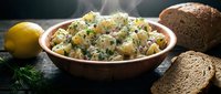 Potato salad with fresh dill and mayonnaise served in a rustic bowl, accompanied by lemon, whole grain bread, and fresh herbs on dark surface.