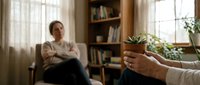 Therapist holding coffee cup during counseling session with patient in cozy office setting with books and plants for mental health support.