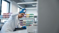 Middle-aged Caucasian male pharmacist in white lab coat working at modern pharmacy counter with colorful medication shelves in background.