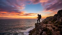 Silhouette of hiker with backpack standing on rocky cliff overlooking ocean at sunset, dramatic sky with orange and pink clouds, adventure and travel concept.