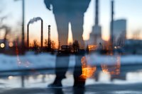 Double exposure of industrial factory silhouettes with smokestacks and urban cityscape reflected in water at sunset, blended with walking person figure.