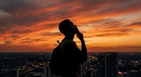Young African American man silhouette using smartphone against dramatic orange sunset sky over city skyline for social media and technology concepts.