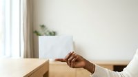 Woman with medium skin tone holding blank white card at wooden table with potted plant and window in background, suitable for business communication, greeting card mockups, and marketing materials.