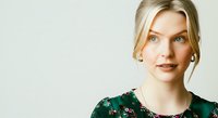 Young woman with blonde hair and fair complexion wearing a dark green floral top, looking at camera with neutral expression against white background for portrait and lifestyle use.