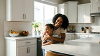Happy African American mother embracing young daughter in bright modern kitchen with white cabinets and natural sunlight streaming through window.