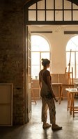 Female artist standing in sunlit studio workspace with easel and art supplies surrounded by brick walls and large windows for creative inspiration.