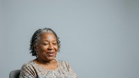 Senior African American woman with gray hair, wearing a beige patterned top, smiling warmly against a neutral gray background. Portrait suitable for healthcare, wellness, diversity, and lifestyle marketing materials.