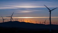 Wind turbines silhouetted against dramatic sunset sky with purple and orange clouds over rolling hills creating renewable energy landscape.