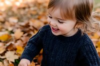 Joyful young girl in navy knit sweater playing with fallen autumn leaves, expressing pure happiness and wonder during fall season outdoor activity.