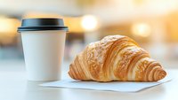 Fresh baked croissant served with takeaway coffee cup on white napkin against blurred cafe background with warm morning light and bokeh effect.
