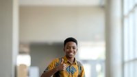 Young man with dark hair and medium brown skin tone wearing a mustard yellow shirt with blue patterned details, smiling confidently in a bright office or modern workspace environment.