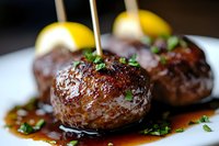 Juicy grilled beef meatballs garnished with fresh herbs and lemon wedges in savory brown sauce on white plate, close-up view of gourmet appetizer.