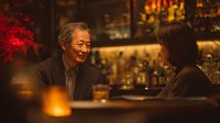 Senior Asian man enjoying conversation with friend at upscale bar counter with warm ambient lighting and bokeh background for hospitality concepts.