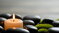 Zen spa composition with burning candle surrounded by smooth black hot stones and green moss creating peaceful meditation atmosphere for wellness.