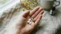 Caucasian woman's hand holding white supplement pills and capsules on neutral linen fabric with dried chamomile flowers, essential oil bottle and ceramic mug nearby for wellness and healthcare content.