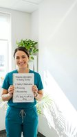 Smiling Hispanic female healthcare worker in teal scrubs holding trauma recovery educational poster in bright medical office with plants for mental health awareness.