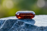 Transparent amber capsule with bubbles on blue stone surface against blurred green background, macro photography showing liquid medicine supplement.