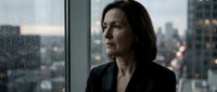 Middle-aged woman with dark hair and serious expression wearing black blazer, standing by window overlooking city skyline, suitable for corporate, business, and professional themes.
