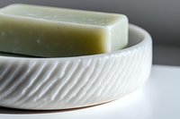 Natural green soap bar in white ceramic dish with diagonal ribbed pattern texture against neutral background, minimalist spa and wellness concept.