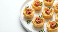 Elegant puff pastry appetizers with prosciutto roses and fresh rosemary garnish on white plate, perfect for holiday parties and gourmet entertaining events.