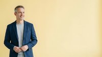 Middle-aged Caucasian male professional wearing navy blazer and light gray shirt, smiling confidently against yellow background. Corporate portrait for business, leadership, and corporate communications.