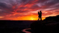 Silhouette of couple embracing on rocky cliff overlooking winding river at dramatic sunset with vibrant orange and red sky, romantic outdoor scene for travel and lifestyle content.