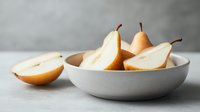 Fresh ripe pears cut in halves arranged in white ceramic bowl on light gray background, minimal food styling for healthy eating concept.