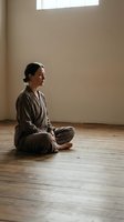 Young Asian woman in comfortable clothing practicing meditation in peaceful minimalist room with natural light for mindfulness and wellness.