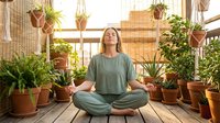 Young woman meditating in lotus position surrounded by potted plants on wooden deck. Peaceful mindfulness practice in urban garden setting with natural light.
