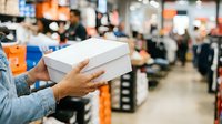 Customer holding white box in warehouse with blurred inventory shelves and staff in background, suitable for retail, logistics, and e-commerce applications.