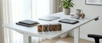 Modern white adjustable desk with wooden letter blocks spelling NOISE, notebooks, coffee mug and plant in bright home office workspace for productivity.