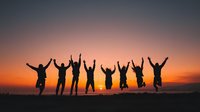 Silhouettes of people jumping with raised arms against dramatic sunset sky, celebrating freedom and achievement together.