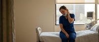 Exhausted healthcare worker in blue scrubs sitting on hospital bed experiencing fatigue and stress during long shift in medical facility.