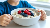 Fresh oatmeal bowl topped with ripe raspberries and blueberries served for breakfast, with child's hands visible at table near window in morning light.
