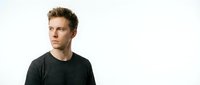 Young man with light brown hair and fair complexion wearing dark charcoal t-shirt, looking thoughtfully to the side against white background for portrait and lifestyle use.