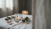 Cozy bedroom scene with lit candles in glass holders and eucalyptus branches on white bedding creating peaceful ambiance for relaxation and wellness.