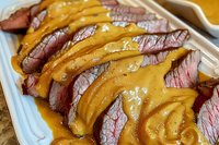 Juicy sliced medium-rare roast beef topped with melted cheese sauce in white container, close-up view showing meat texture and glistening sauce drizzle.