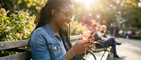 Young African American woman with dreadlocks smiling while using smartphone on park bench during golden hour sunset with warm natural lighting and bokeh background.