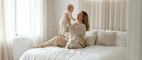 Young mother holding up smiling baby in bright white bedroom with natural light streaming through windows creating warm intimate family moment.