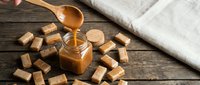Homemade salted caramel sauce dripping from wooden spoon into glass jar surrounded by golden caramel candies on rustic wood table for baking recipes.
