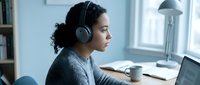 Woman with curly dark hair and medium skin tone wearing wireless headphones, sitting at desk with laptop, mug, and lamp. Remote work, online learning, or virtual communication concept.