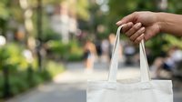 Person with medium skin tone holding white canvas tote bag outdoors in urban park setting, suitable for retail, shopping, sustainability and eco-friendly lifestyle marketing campaigns.